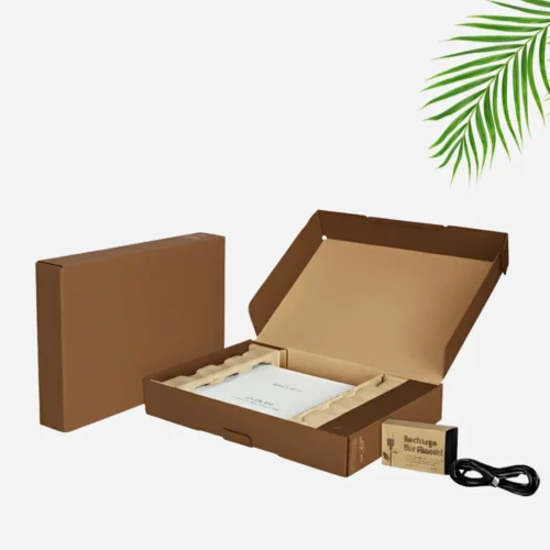 An open brown cardboard mailer box reveals a laptop (partially obscured by a white protective cover) and brown paper-based inserts. Next to it, a closed brown cardboard box is visible. In the foreground, a small brown box with "Recharge Our Planet!" printed on it and a coiled black power cable are shown.