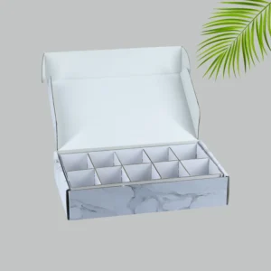 A close-up shot of a white and gray marble-patterned mailer box with its lid open. Inside, a cardboard divider creates 12 separate compartments. The interior of the box and the divider are plain white.