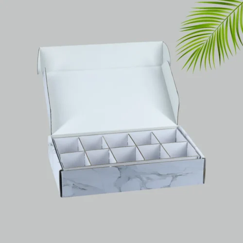 A close-up shot of a white and gray marble-patterned mailer box with its lid open. Inside, a cardboard divider creates 12 separate compartments. The interior of the box and the divider are plain white.
