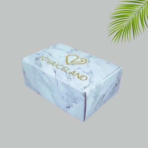 A closed rectangular mailer box with a white marble design. The lid displays a gold heart-shaped logo above the brand name in bold gold lettering.