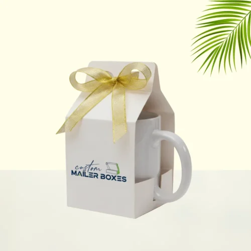 A white mug gift box with a "Custom Mailer Boxes" logo printed on the front. The box is tied with a gold ribbon bow, and the mug handle is visible on the side.