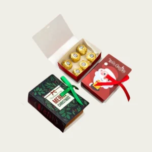 A collection of Christmas sweet packaging boxes shaped like small books. One is dark green with a holly design and a green ribbon, and the other is red with a Santa image and a red ribbon. A small white box insert holds six foil-wrapped chocolates.