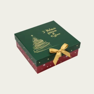 A luxury two-piece Christmas gift box with a dark green lid, gold foil script reading Happy New Year and Merry Christmas, a sketch of a Christmas tree, and a red base with white and gold snowflakes, tied with a gold satin bow.